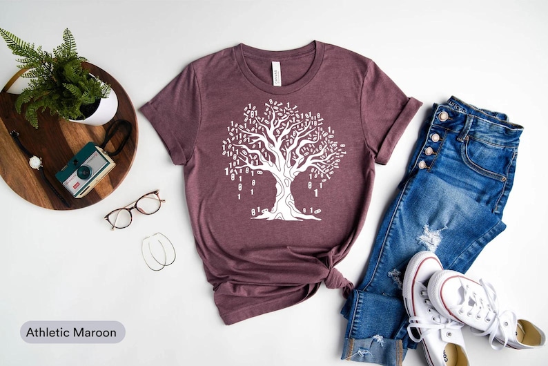 A casual outfit consisting of a maroon t-shirt with a white tree design, blue jeans, and white sneakers, along with a pair of glasses and a potted plant, is laid out on a white background.