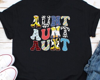 May include: Black t-shirt featuring the word "AUNT" repeated three times in a patchwork design. Each letter is styled as a cowboy boot with various patterns, including cow print, plaid, and floral prints. The shirt is paired with jeans and white trainers.