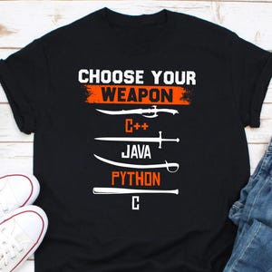 Puede incluir: Camiseta negra con el texto "CHOOSE YOUR WEAPON" en blanco y naranja. Debajo, ilustraciones de espadas representan C++, Java, Python y C. La camiseta se combina con zapatillas blancas y vaqueros azules.