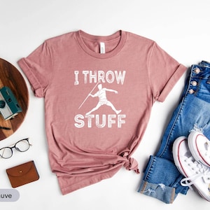 Puede incluir: Camiseta color malva con la frase "I Throw Stuff" y un gráfico de una persona lanzando una jabalina. La camiseta se combina con vaqueros azules y zapatillas blancas. Otros accesorios incluyen una bandeja de madera, gafas y una cámara.