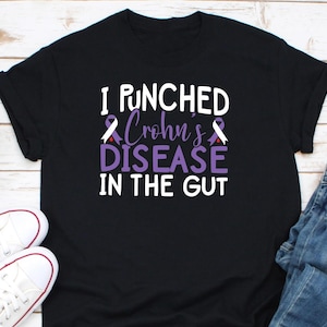 Op de afbeelding: Zwart T-shirt met de tekst "I PUNCHED Crohn's DISEASE IN THE GUT" in wit en paars. Het ontwerp bevat paarse bewustzijns linten. Witte sneakers en een blauwe jeans zijn zichtbaar.