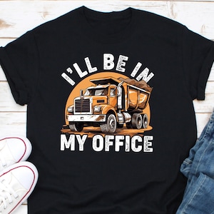 May include: Black t-shirt featuring a cartoon dump truck design with the text "I'll Be In My Office" in white. The truck is orange and brown, set against a yellow sun-like background.