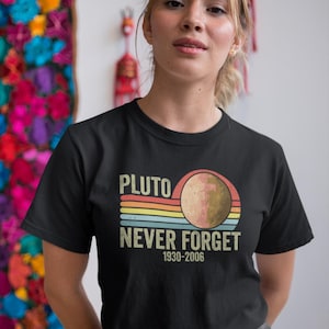 May include: Black t-shirt with a retro design featuring the text "PLUTO NEVER FORGET 1930-2006" and an illustration of Pluto. The design includes a planet graphic with a rainbow-colored stripe background.