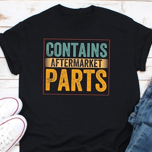 Könnte beinhalten: Schwarzes T-Shirt mit dem Text "CONTAINS AFTERMARKET PARTS" in Türkis, Gelb und Orange. Der Text befindet sich in einem rechteckigen Rahmen. Weiße Turnschuhe und blaue Jeans sind im Bild zu sehen.