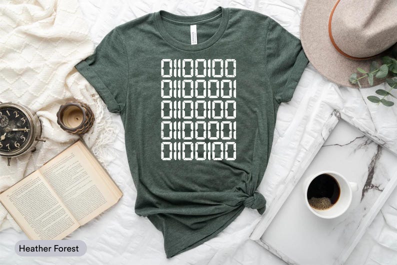a green t-shirt with a white digital design, a hat, a book, a cup of coffee, and a clock, all arranged on a bed.