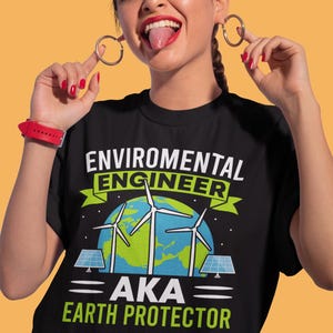 Puede incluir: Camiseta negra con el texto "ENVIRONMENTAL ENGINEER AKA EARTH PROTECTOR" con un gráfico de aerogeneradores, paneles solares y un globo terráqueo. La persona lleva pendientes de aro y un reloj rojo.