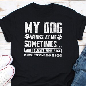 May include: Black t-shirt with white text that reads "MY DOG WINKS AT ME SOMETIMES... AND I ALWAYS WINK BACK IN CASE IT'S SOME KIND OF CODE!". The design includes paw prints. A pair of white sneakers is visible.