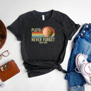 May include: Dark heather gray t-shirt with the text "PLUTO NEVER FORGET 1930-2006" and a graphic of Pluto. The shirt is paired with blue jeans and white sneakers. Other accessories include a brown wallet, glasses, and a camera.