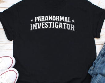 May include: Black t-shirt with white text that reads "Paranormal Investigator" with a star on either side of the text.
