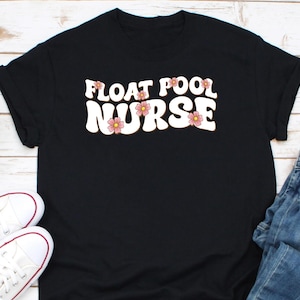 May include: Black t-shirt with the words "FLOAT POOL NURSE" in a retro font, decorated with pink flowers. The shirt is paired with white sneakers and blue jeans, suggesting a casual, everyday style.