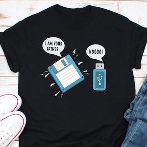 Pode incluir: Uma camiseta preta com um gráfico de um disquete e uma unidade USB. O disquete diz "Eu sou seu pai" e a unidade USB diz "Noooo!"