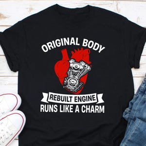 May include: Black t-shirt with a red heart and a motorcycle engine graphic. The text "Original Body, Rebuilt Engine, Runs Like A Charm" is printed on the shirt.