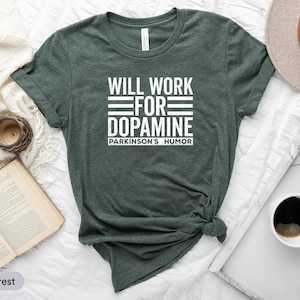 Op de afbeelding: Een donkergroen t-shirt met de witte tekst "WILL WORK FOR DOPAMINE" en "PARKINSON'S HUMOR" eronder. Het shirt wordt gepresenteerd op een wit oppervlak met een boek, een klok, een kandelaar, een hoed en een kopje koffie.