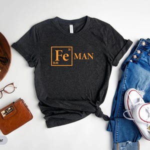 a casual outfit consisting of a black t-shirt with a chemical element design, a pair of blue jeans, white sneakers, and a brown wallet. The items are arranged on a white background, creating a visually appealing and organized composition.