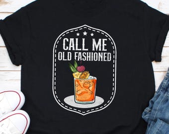 May include: Black t-shirt featuring a white graphic with the text "CALL ME OLD FASHIONED." The graphic includes an illustration of an Old Fashioned cocktail with a cherry and pineapple garnish. The shirt is paired with white sneakers and blue jeans.