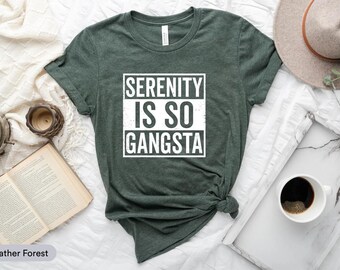May include: A heather forest green t-shirt with the text "SERENITY IS SO GANGSTA" in a white, rectangular design. The shirt is styled with a knot at the bottom. Other items in the image include a book, a clock, and a coffee cup.