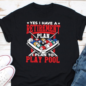 May include: Black t-shirt with the text "YES I HAVE A RETIREMENT PLAN I PLAN TO PLAY POOL" in red and white. The design includes a rack of billiard balls and crossed pool cues.