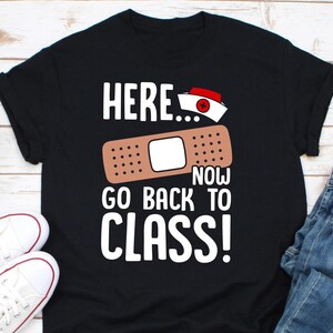 Op de afbeelding: Zwart T-shirt met witte tekst die "Here... Now Go Back To Class!" luidt en een cartoon afbeelding van een pleister met een rood kruis.