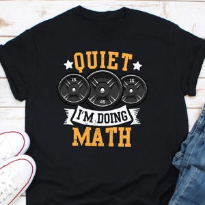 以下が含まれることがあります： 「Quiet I'm doing math」というテキストと、ウェイトプレートのグラフィックが描かれた黒いTシャツ。