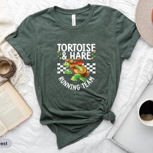 May include: Heather forest green t-shirt featuring a cartoon design of a tortoise and a hare racing. The text "Tortoise & Hare Running Team" is printed above and below the graphic. The shirt is made of a soft material.