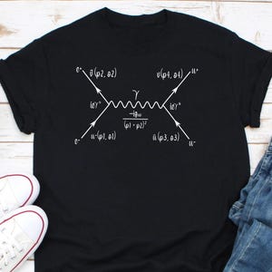 May include: A black t-shirt with a white graphic of a Feynman diagram, a representation of a particle interaction in physics. The diagram shows an electron and a positron interacting via a photon, with the text '-igμν/(p1 + p2)2' above the photon line.