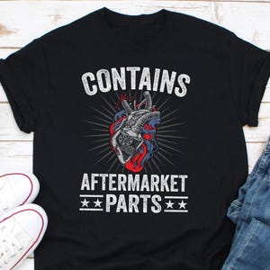 Könnte beinhalten: Schwarzes T-Shirt mit einem Grafikdruck eines mechanischen Herzens und dem Text "CONTAINS AFTERMARKET PARTS" in Weiß.