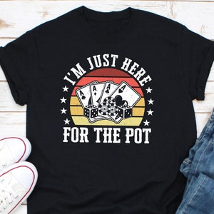 May include: Black t-shirt with a poker-themed graphic. The design features playing cards, dice, and poker chips set against a sunset background. The text reads "I'M JUST HERE FOR THE POT."