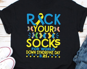 Pode incluir: Camiseta preta com o texto "Rock Your Socks" em azul e amarelo, com uma fita amarela e um gráfico de meias coloridas. A camiseta promove o Dia Mundial da Síndrome de Down, com a data 3.21.