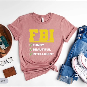 Op de afbeelding: Heather mauve t-shirt met de gele tekst "FBI" en de woorden "Funny, Beautiful, Intelligent" eronder. De afbeelding bevat een blauwe spijkerbroek en witte sneakers.