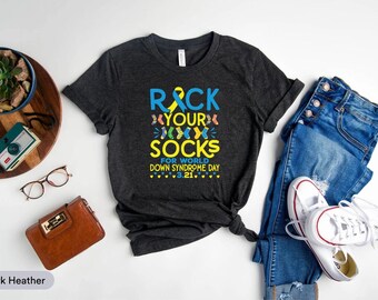 Pode incluir: T-shirt cinzento escuro com o texto "Rock Your Socks for World Down Syndrome Day 3.21." A camisa é combinada com jeans azuis e tênis brancos. Outros acessórios incluem uma carteira castanha e óculos.
