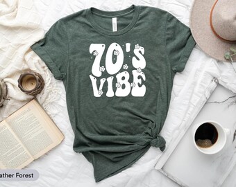 Puede incluir: Camiseta verde bosque jaspeado con el texto blanco "70's Vibe" y gráficos de flores. La camiseta está anudada en el dobladillo. Un sombrero, una taza de café y un libro abierto están en el fondo. La camiseta es de material suave.