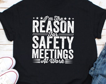 May include: Black t-shirt with white text that reads "I'm The REASON For SAFETY MEETINGS At Work." The text is in a bold, blocky font. The shirt is folded, with white sneakers and blue jeans visible.