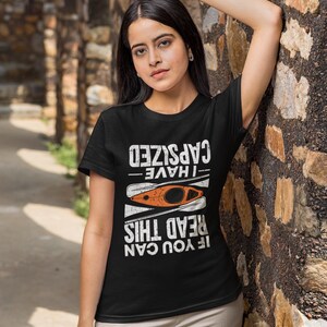 May include: Black t-shirt with the text "I HAVE CAPSIZED IF YOU CAN READ THIS" in white. An orange kayak graphic is centered below the text. The shirt is worn by a person.