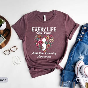 May include: Athletic maroon t-shirt with the text "EVERY LIFE Has Value" and "Addiction Recovery Awareness" above a floral heart with a semicolon. The shirt is paired with blue jeans and white sneakers.