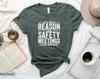May include: A heather forest green t-shirt with the text "I'm The Reason For Safety Meetings At Work" in white. The shirt is styled with a knot at the bottom. A hat, coffee cup, and open book are in the background.