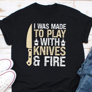 Op de afbeelding: Zwart T-shirt met een witte afbeelding waarop staat "I was made to play with knives & fire". De afbeelding bevat ook een mes en twee flessen.