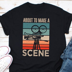 Könnte beinhalten: Schwarzes T-Shirt mit einer Grafik im Vintage-Stil einer Filmkamera und dem Text "About to make a scene".