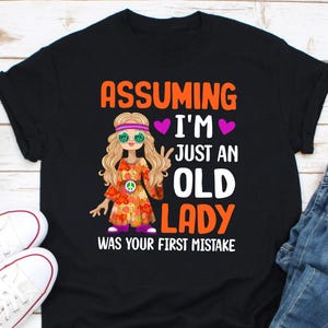 Puede incluir: Camiseta negra con la frase "Assuming I'm Just An Old Lady Was Your First Mistake". La camiseta presenta una caricatura de una mujer con ropa hippie, gafas de sol y un símbolo de la paz. La mujer lleva un vestido floral.