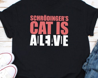 May include: Black t-shirt with the text "Schrödinger's Cat Is Alive" in a distressed font. The text is in pink and white. The shirt is displayed on a light wood surface with white sneakers and blue jeans.