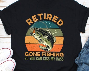Puede incluir: Camiseta negra con un diseño gráfico de un róbalo saltando. El texto "RETIRED GONE FISHING SO YOU CAN KISS MY BASS" está en un estilo vintage con un fondo de puesta de sol.
