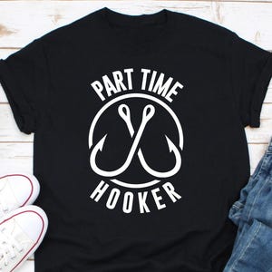 May include: Black t-shirt with the text "PART TIME HOOKER" in a white, circular design featuring two crossed fishing hooks. The shirt is paired with blue jeans and white sneakers.