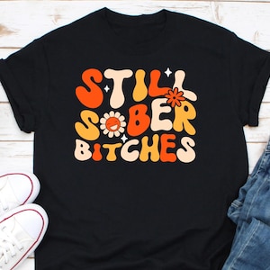 May include: Black t-shirt with the text "STILL SOBER BITCHES" in orange, yellow, and white retro font. The design includes a smiling flower and star accents. White sneakers and blue jeans are visible in the image.