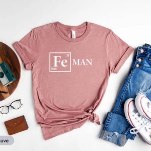 a pink t-shirt with the text "Fe Man" printed on it, along with a pair of jeans and sneakers, as well as various accessories such as a wallet, glasses, and a camera.