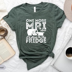 Może przedstawiać: Zielona koszulka w kolorze wrzosu z napisem "ONE MORE MRI And I'll Stick To The Fridge" w kolorze białym. Koszulka przedstawia grafikę rolki ręczników papierowych. Koszulka jest prezentowana na białej powierzchni z książką, zegarem i filiżanką kawy.