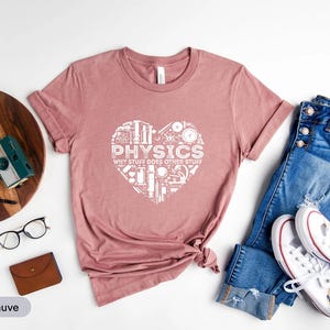 May include: Heather mauve t-shirt with a white heart-shaped graphic containing physics-related illustrations and the text "PHYSICS WHY STUFF DOES OTHER STUFF". The shirt is paired with blue jeans and white trainers.