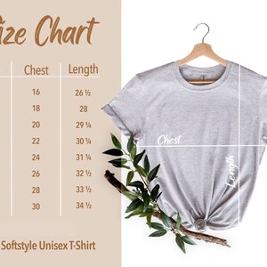 May include: A light gray Adult Softstyle Unisex T-Shirt hangs on a wooden hanger. A size chart shows chest and length measurements in inches for sizes XS to 4XL. The chart is next to the shirt.