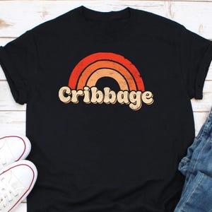 May include: Black t-shirt featuring a retro rainbow design in shades of red, orange, and beige. The word "Cribbage" is printed in a vintage font below the rainbow. The shirt is paired with white sneakers and blue jeans.