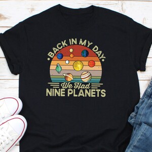 May include: Black t-shirt with a retro planet design. The text reads "BACK IN MY DAY. WE HAD NINE PLANETS." The design features a sunset background and various planets. A fun, vintage-style tee.
