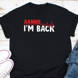 May include: Black t-shirt with the text "AANND... I'M BACK" in white and red. The design includes a red heartbeat line graphic. The shirt is paired with white trainers and blue jeans.