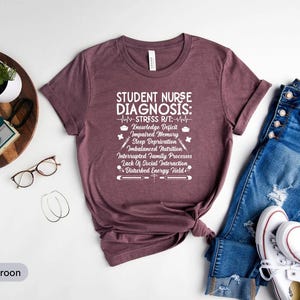 May include: Athletic maroon t-shirt with the text "STUDENT NURSE DIAGNOSIS" and a list of potential issues. The shirt is paired with blue jeans and white sneakers. A watch, glasses, and a camera are also in the image.